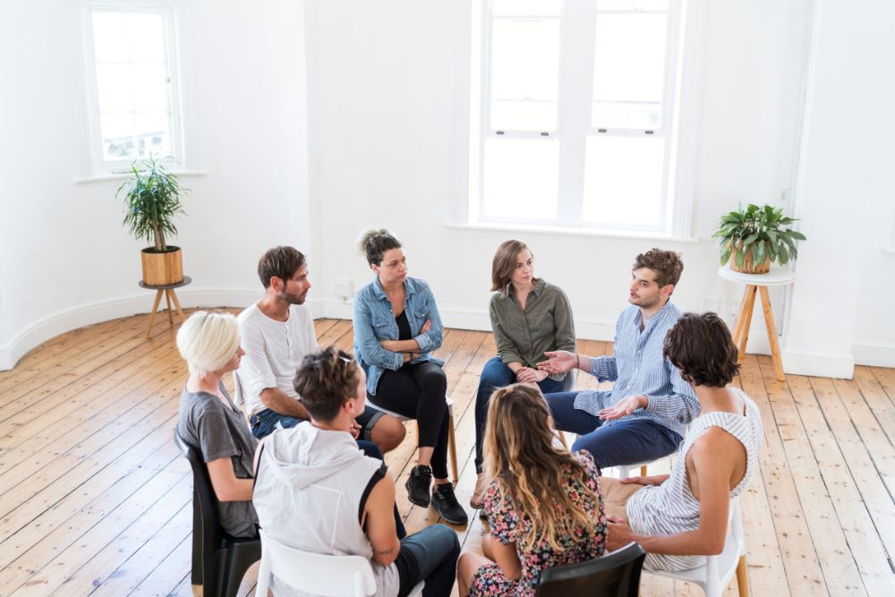 Personengruppe in einem hellen Raum bei einer Gruppentherapie, die in einer Gesprächsrunde zusammenkommt.