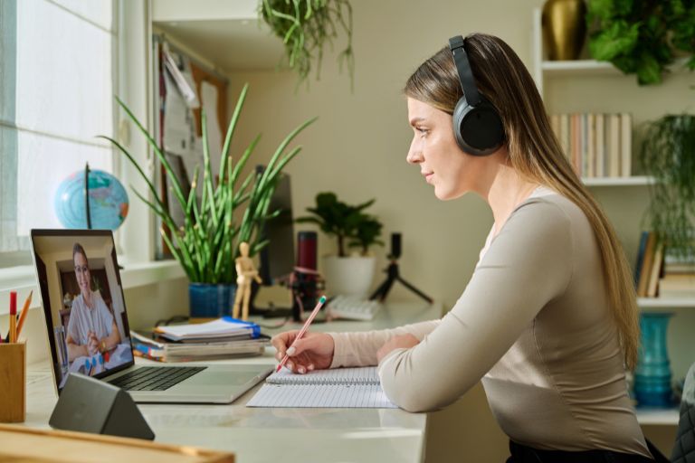 Eine Frau mit Kopfhörern macht Notizen während einer Online-Psychotherapie-Sitzung am Laptop.
