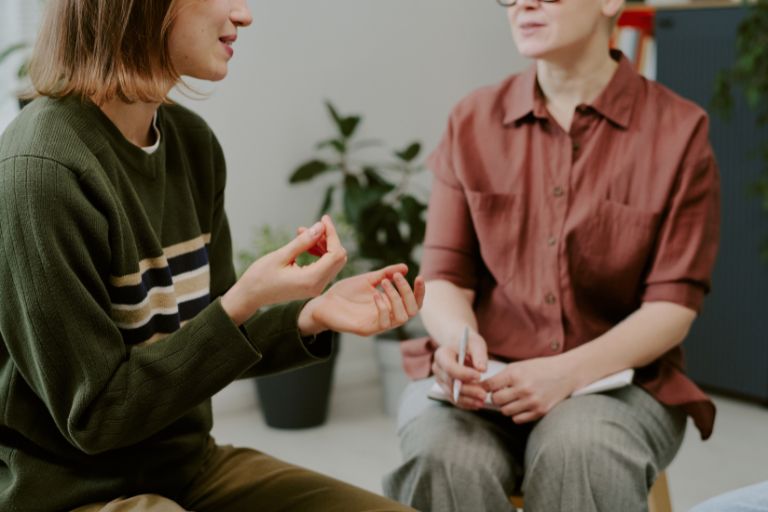Zwei Frauen im Gespräch bei einer psychosozialen Beratung, eine spricht gestikulierend, die andere macht sich Notizen.